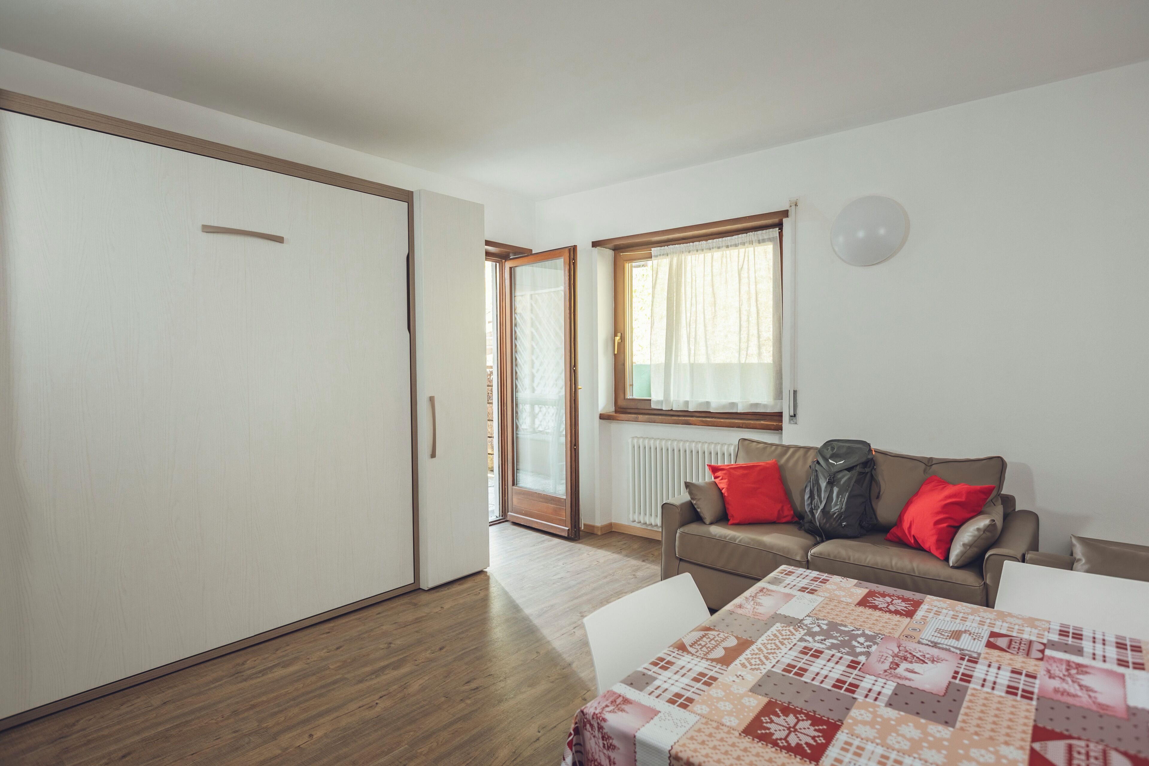 A room with a sliding door, a sofa with red cushions, a table with a patterned tablecloth, and a window with curtains.