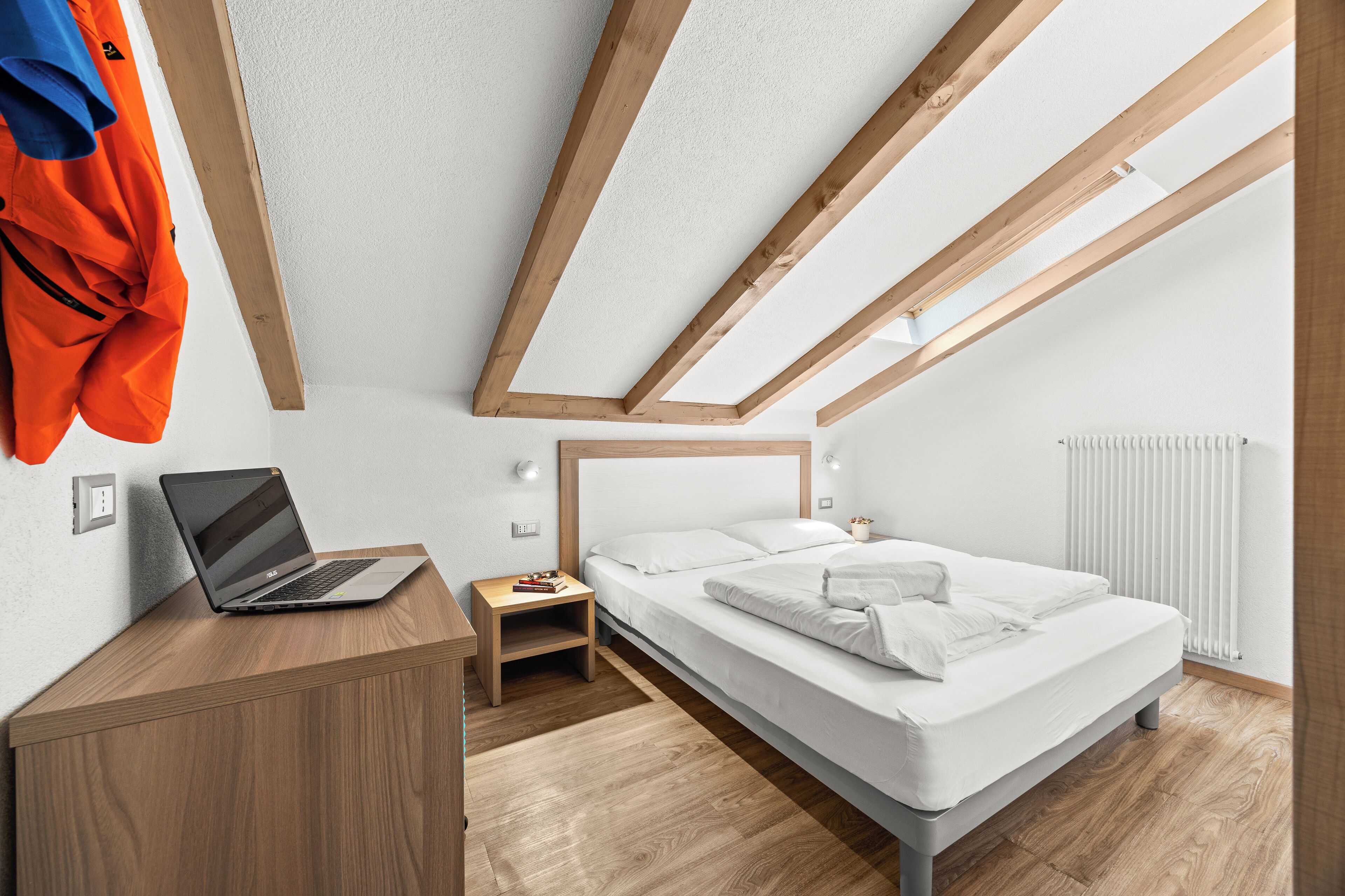 A modern bedroom with a sloped ceiling, a bed, a wooden desk with a laptop, and a radiator.