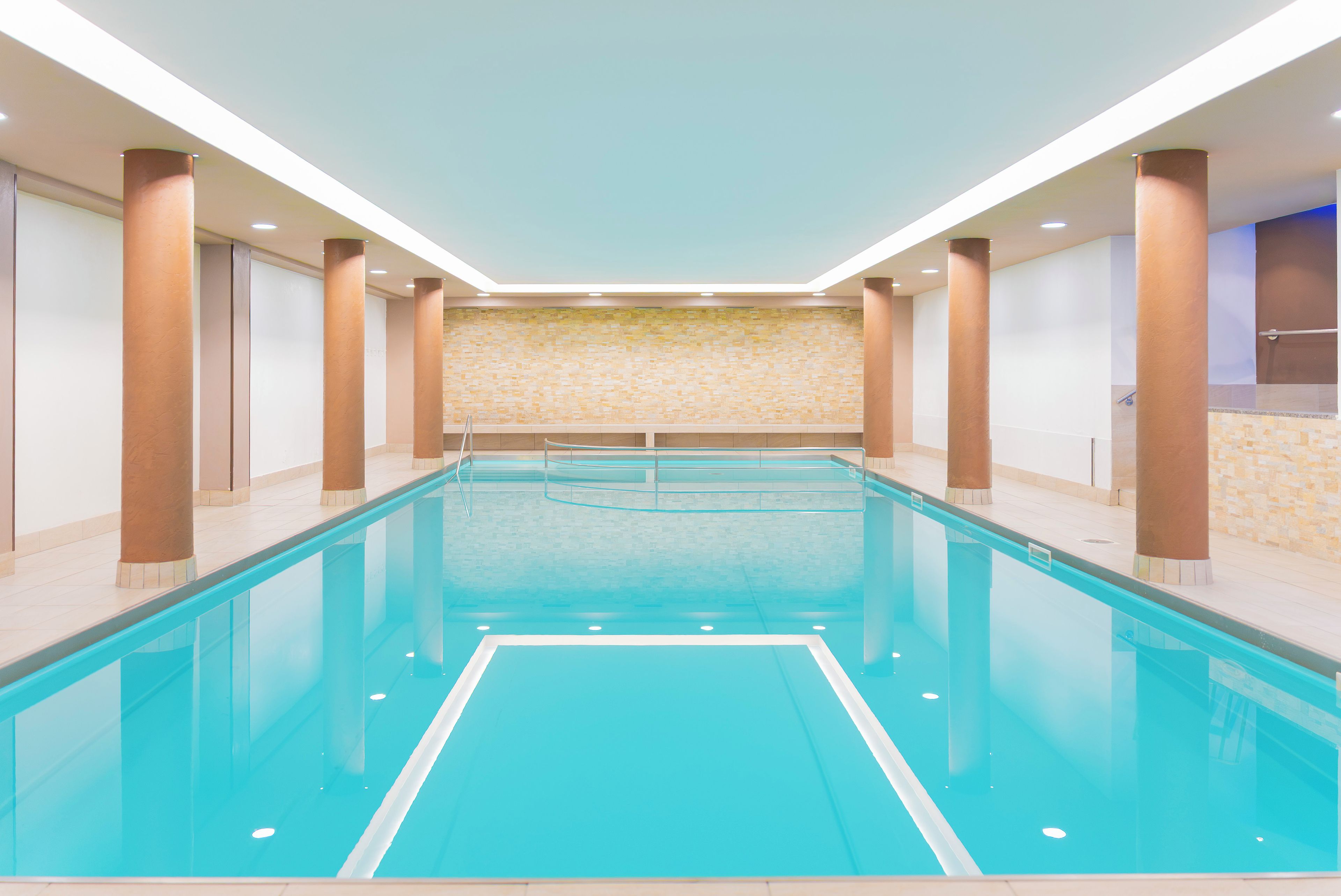 Une longue et étroite piscine intérieure, avec une eau d’un bleu limpide, entourée de murs à motifs et de grandes colonnes.