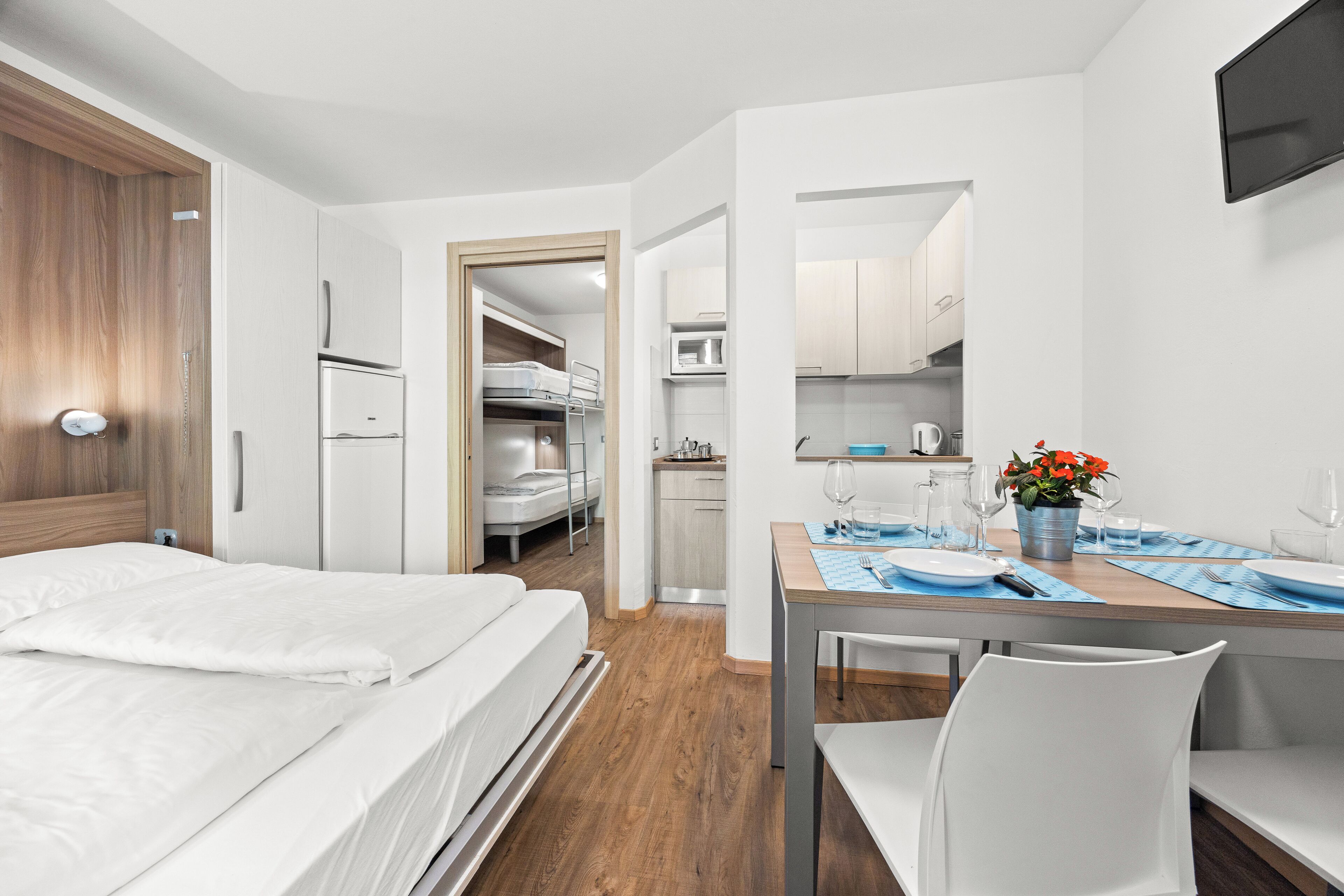 A hotel room with a bed, a dining table with chairs, a kitchen area, and a TV mounted on the wall.