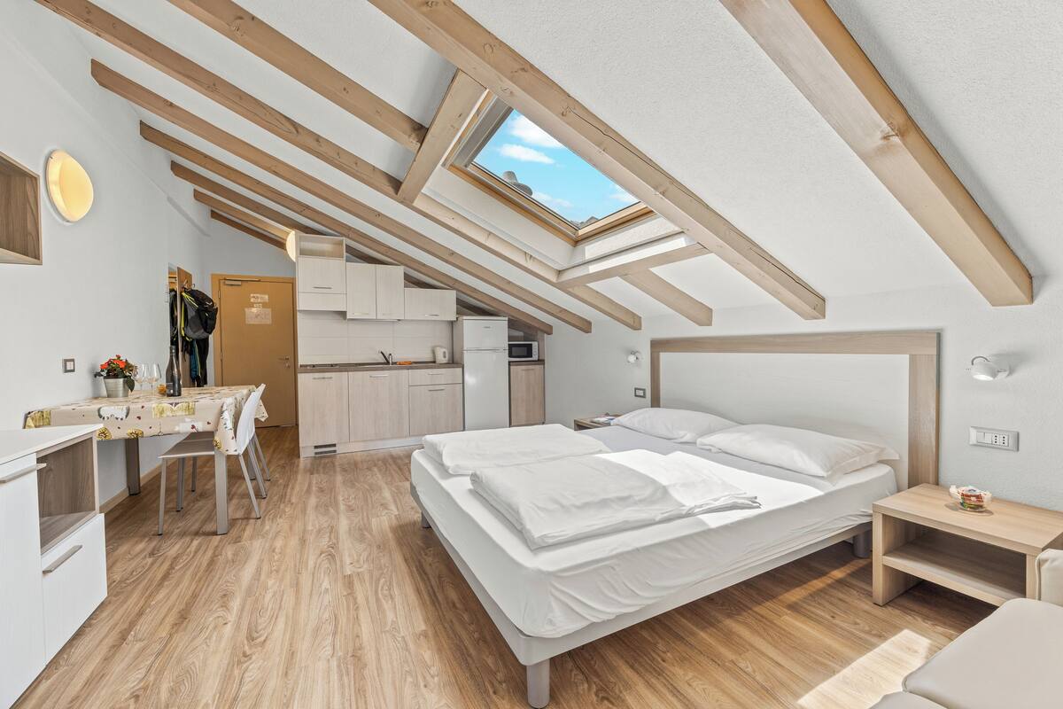 A modern bedroom with a skylight, wooden beams, a bed, a desk, and a kitchenette.