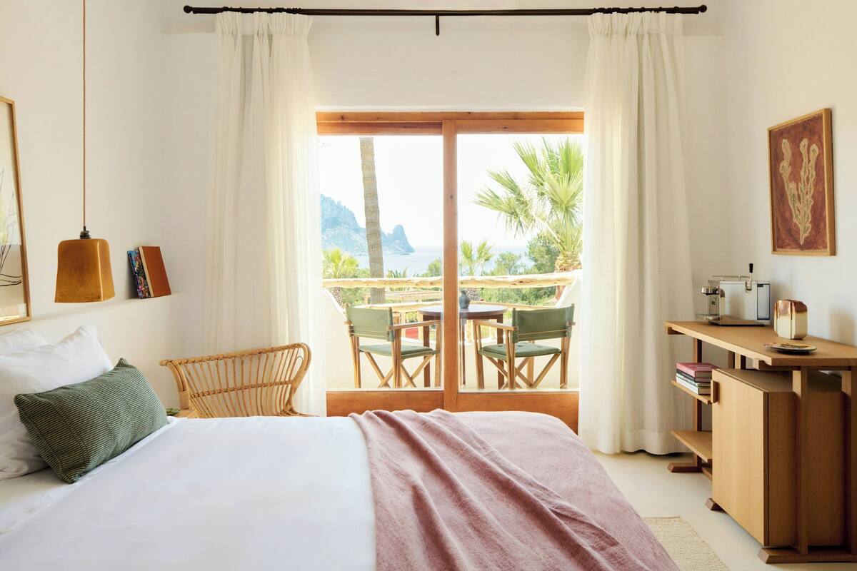 A bedroom with a bed, a desk, a chair, and a view of the outdoors.