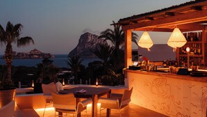 An outdoor dining area with a view of the sea and a mountain, featuring a bar with a wooden counter and pendant lights. - Petunia Ibiza, a Beaumier Hotel (Sant Josep de sa Talaia)