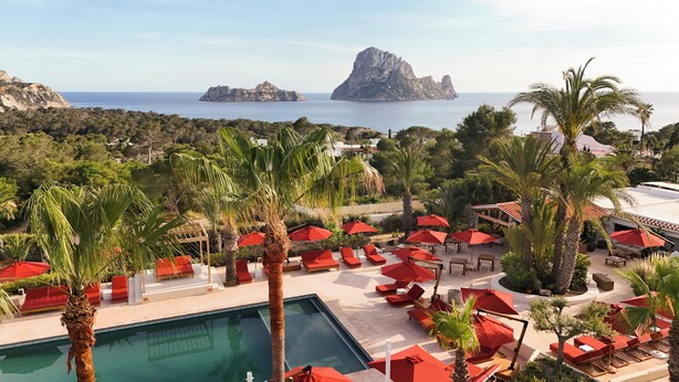 A pool area with red umbrellas and lounge chairs, surrounded by palm trees and overlooking the sea. - Petunia Ibiza, a Beaumier Hotel (Sant Josep de sa Talaia)