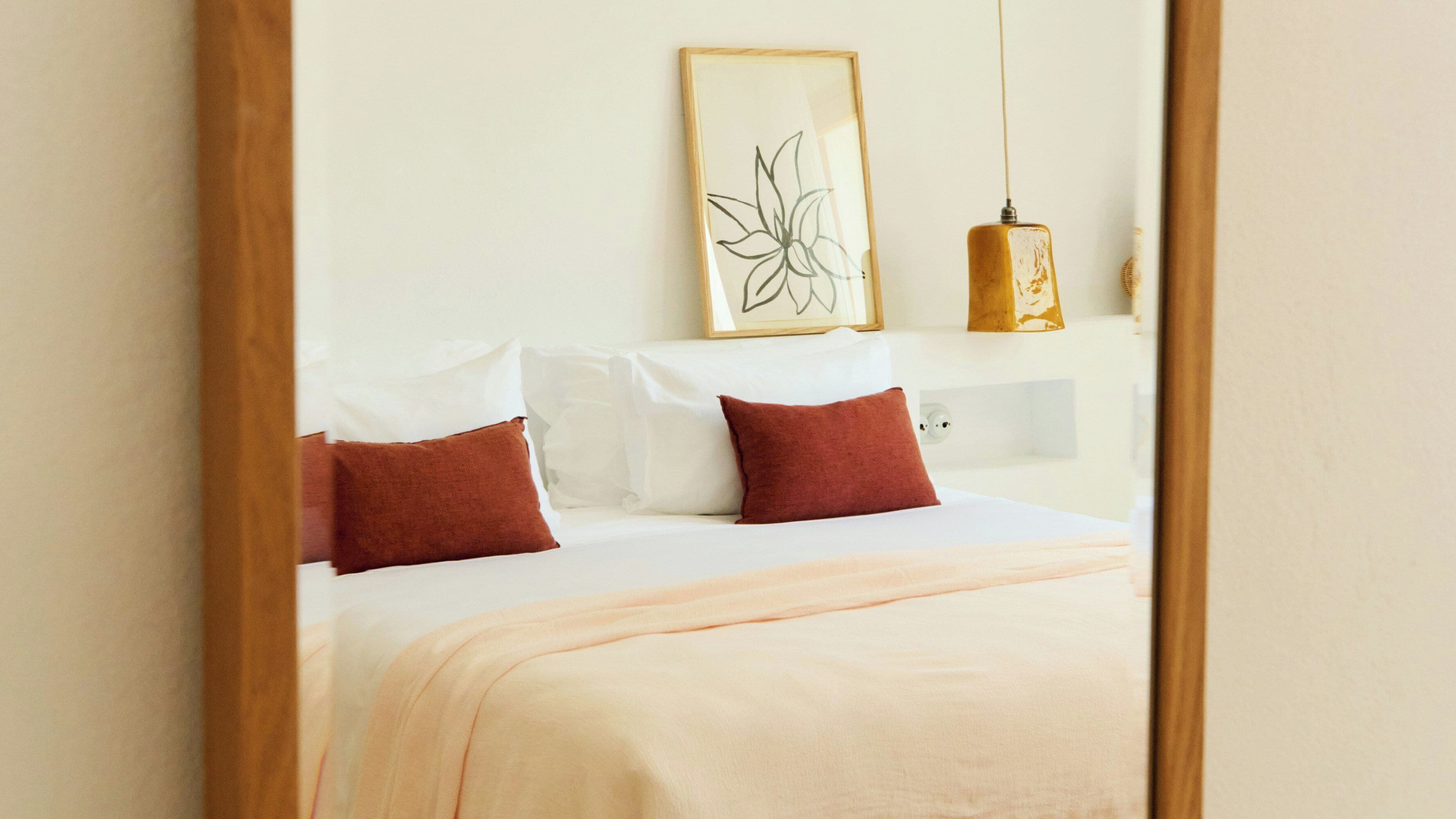 A neatly made bed with white and red pillows, a beige blanket, and a framed floral artwork above it.