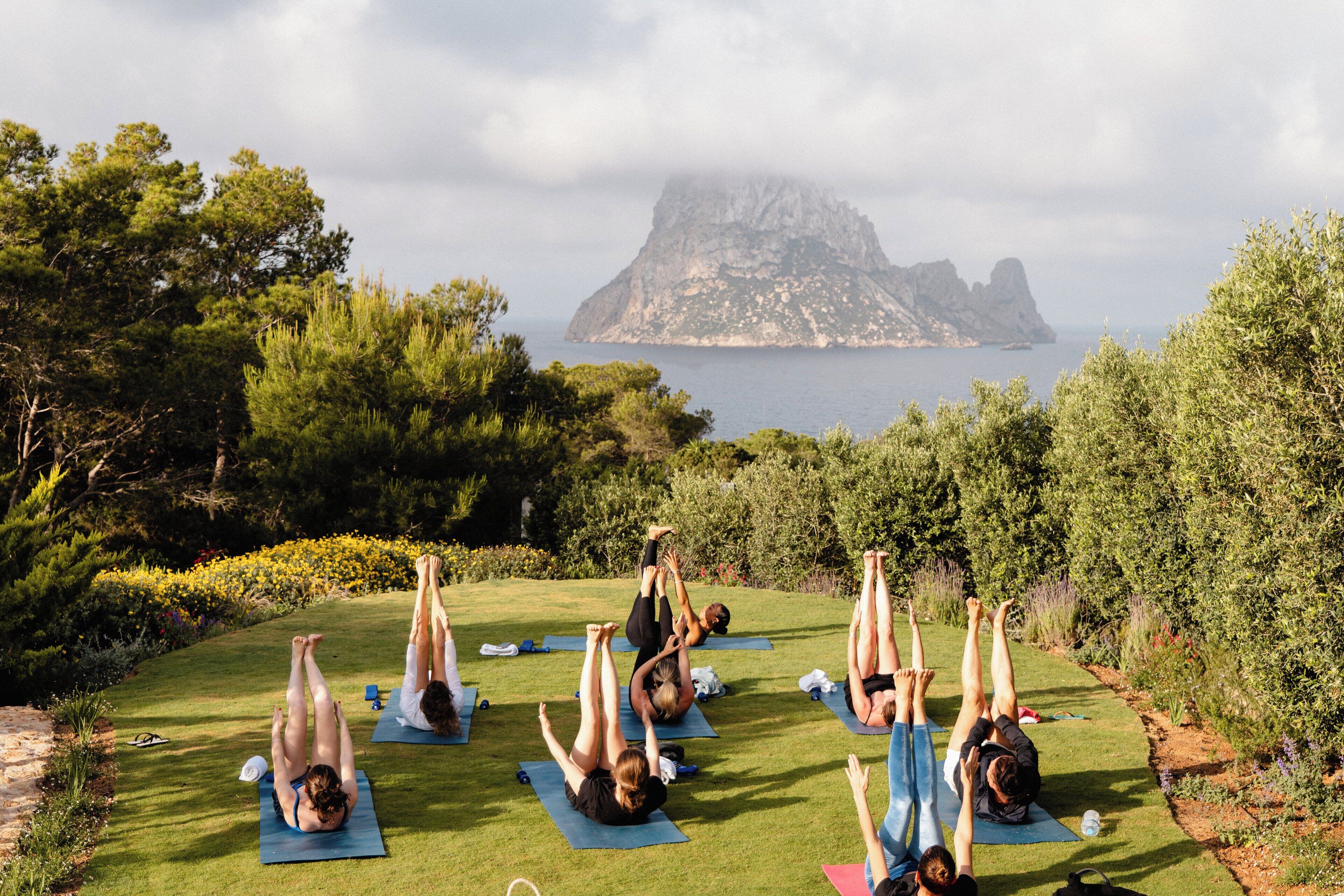 A group of people doing yoga on mats in a grassy area with a view of a rocky island in the distance.