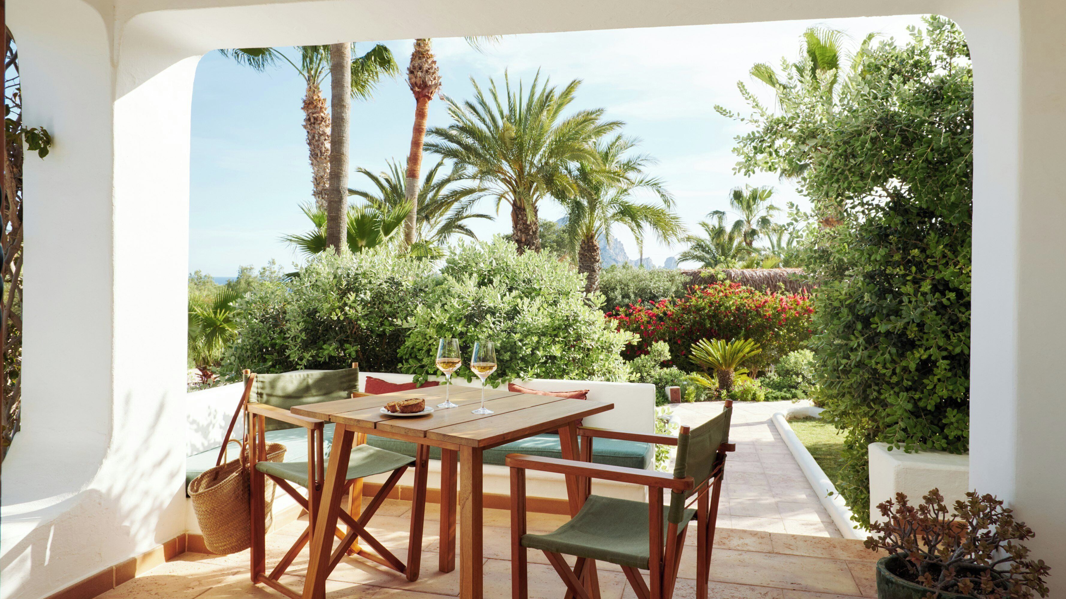 A patio with a wooden table and chairs, a woven basket, and a view of lush greenery and palm trees.
