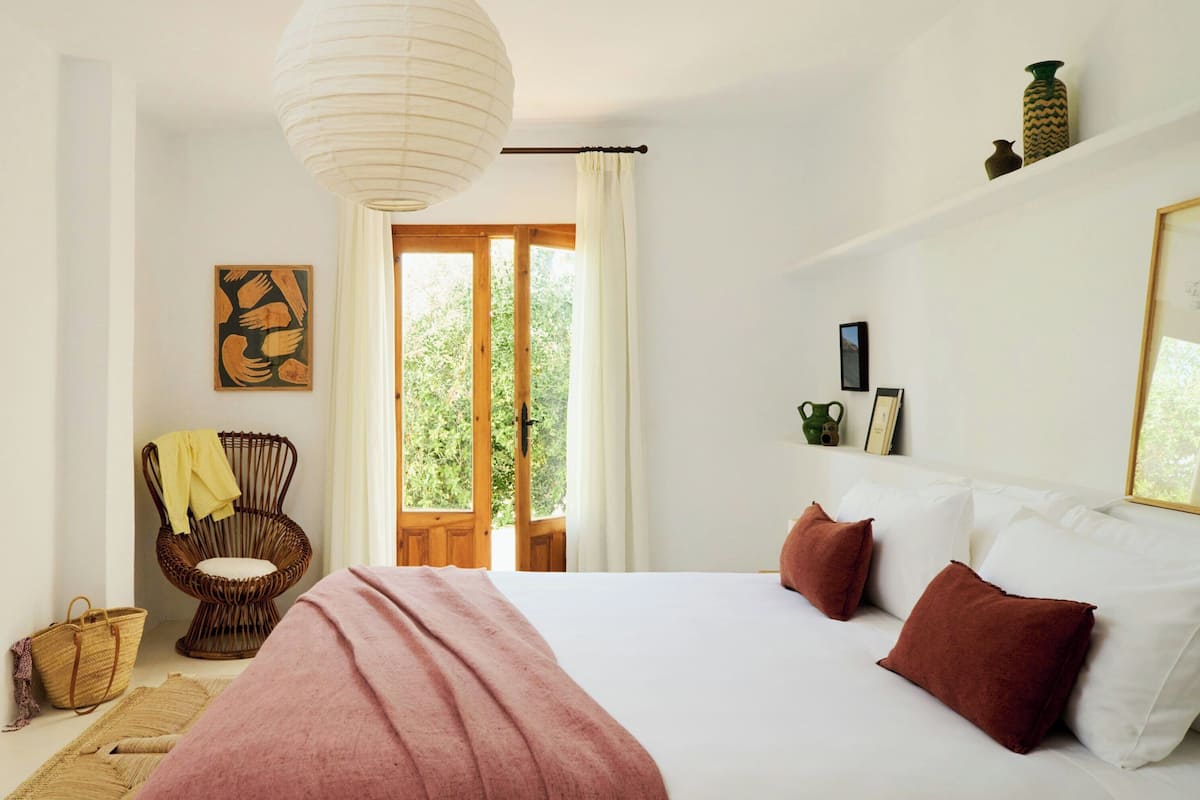 A bedroom with a bed, a wicker chair, a basket, a painting, and a view through a door.