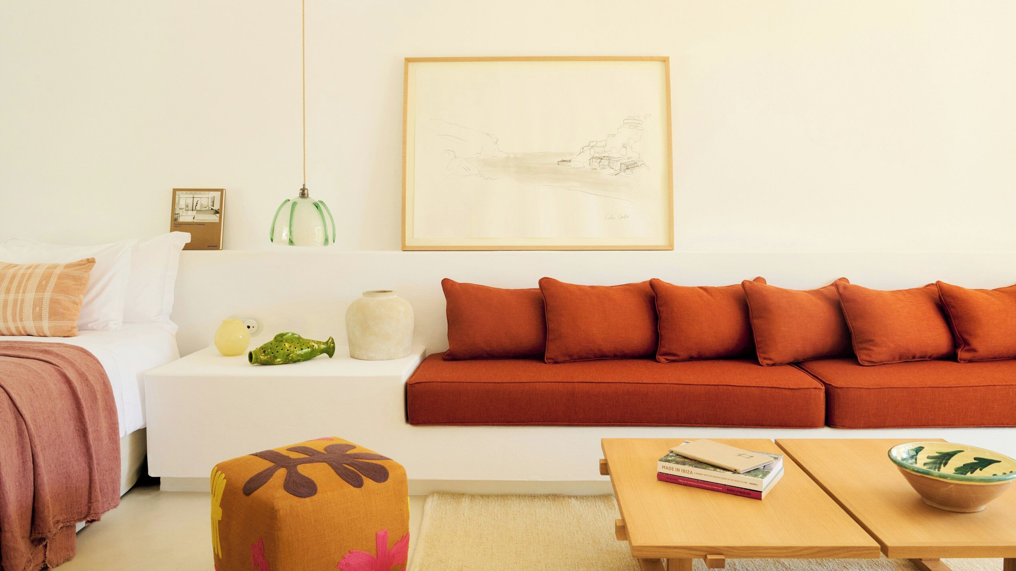 A living room with a white sofa, orange cushions, a small table with books, and a green vase.
