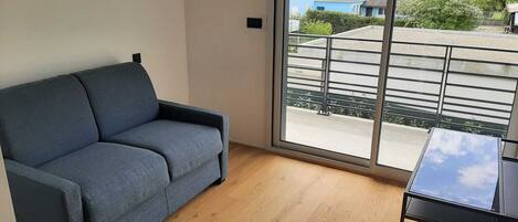 Ein modernes Wohnzimmer mit einer grauen Couch, einem kleinen schwarzen Tisch und einem großen Fenster mit Blick auf eine Wohngegend.