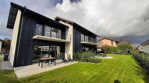 A modern two-story house with a large balcony, a well-maintained lawn, and a dining area with outdoor furniture.