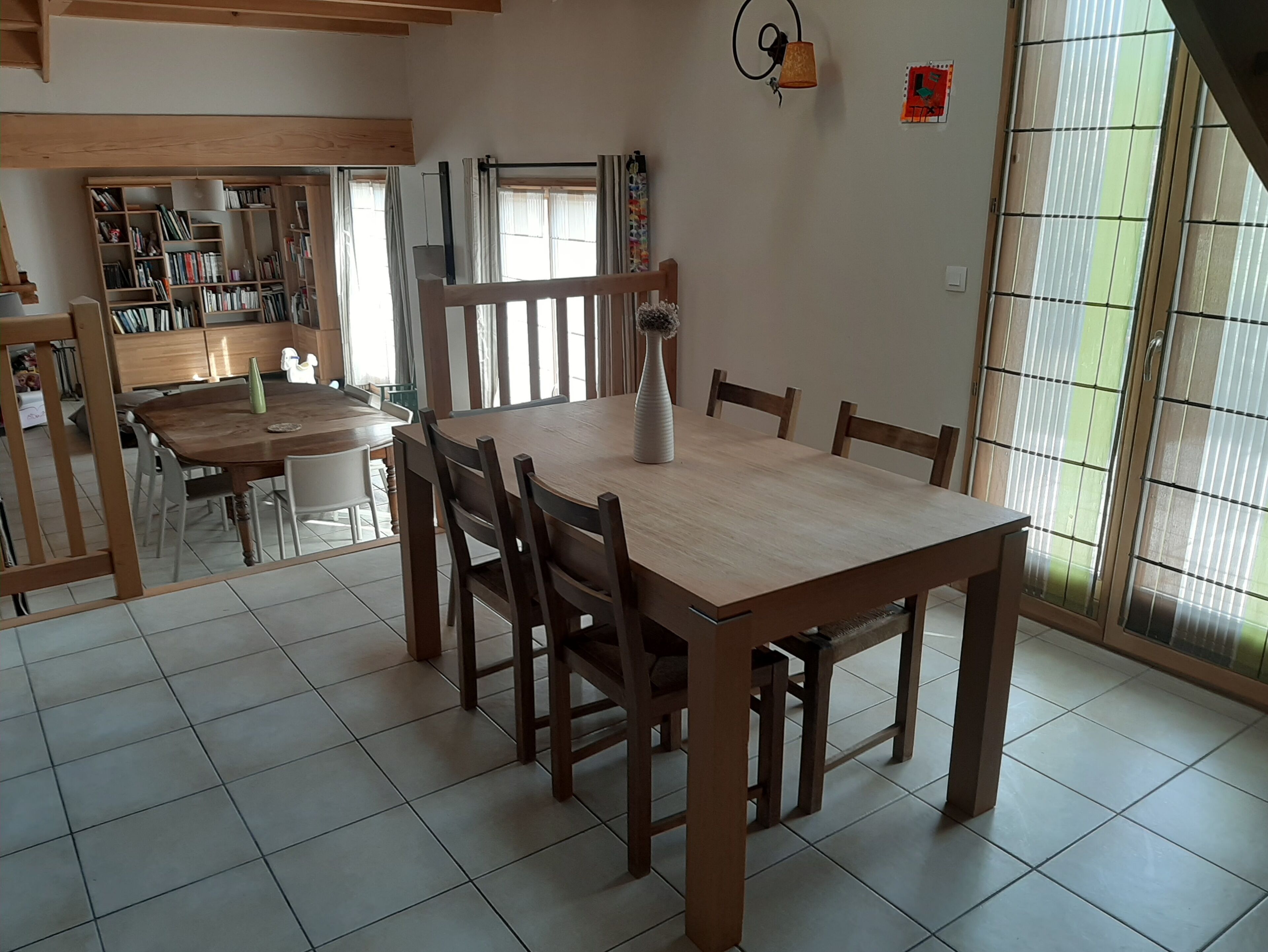 A dining area with a wooden table and chairs, a bookshelf, and a window with curtains.