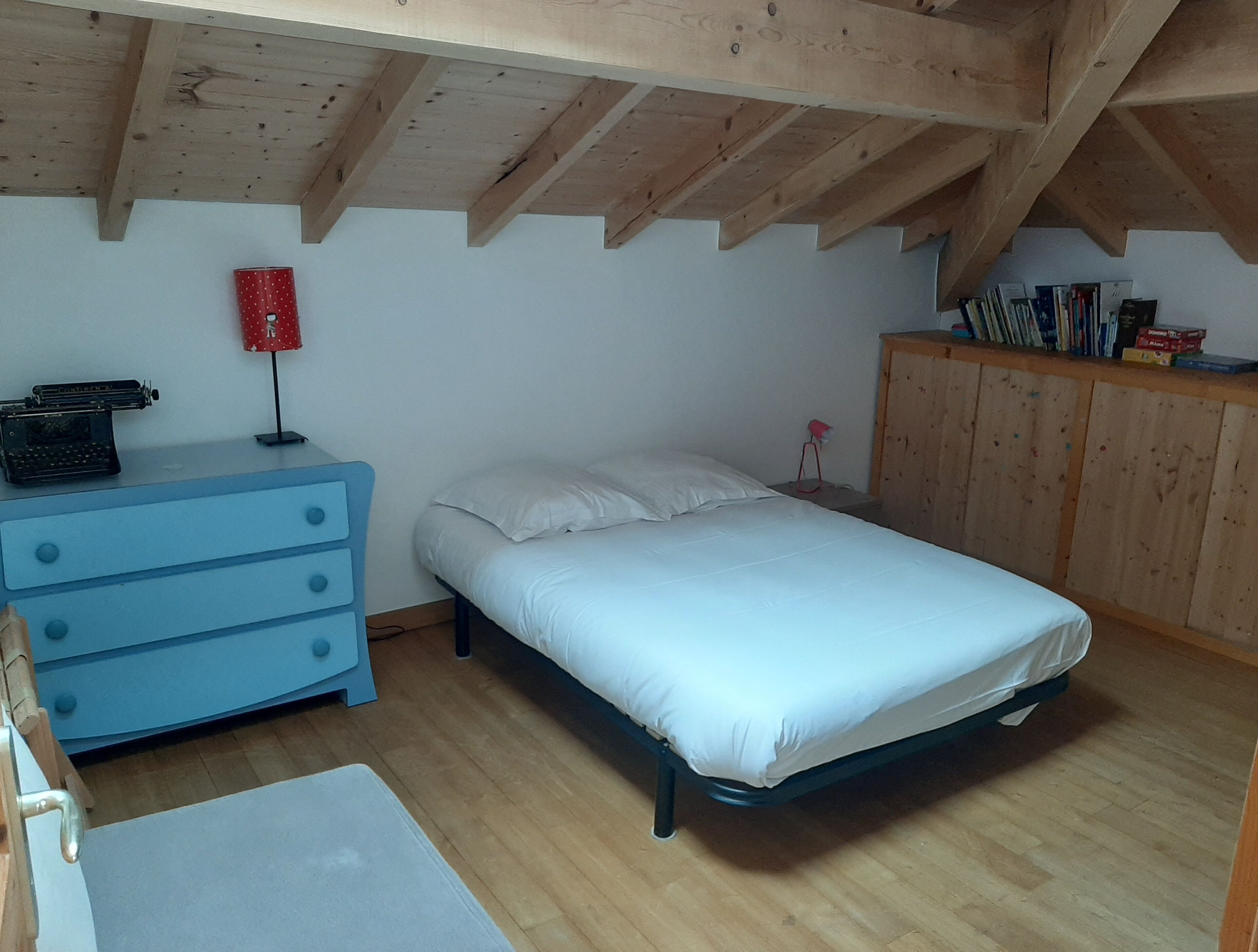 A bedroom with a wooden ceiling, a bed, a dresser, and a small desk with a typewriter.