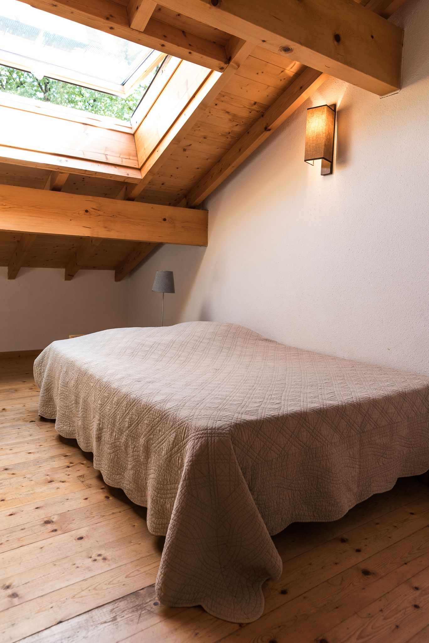 A bedroom with a sloped ceiling, a skylight, a bed with a beige blanket, and a wall-mounted light fixture.