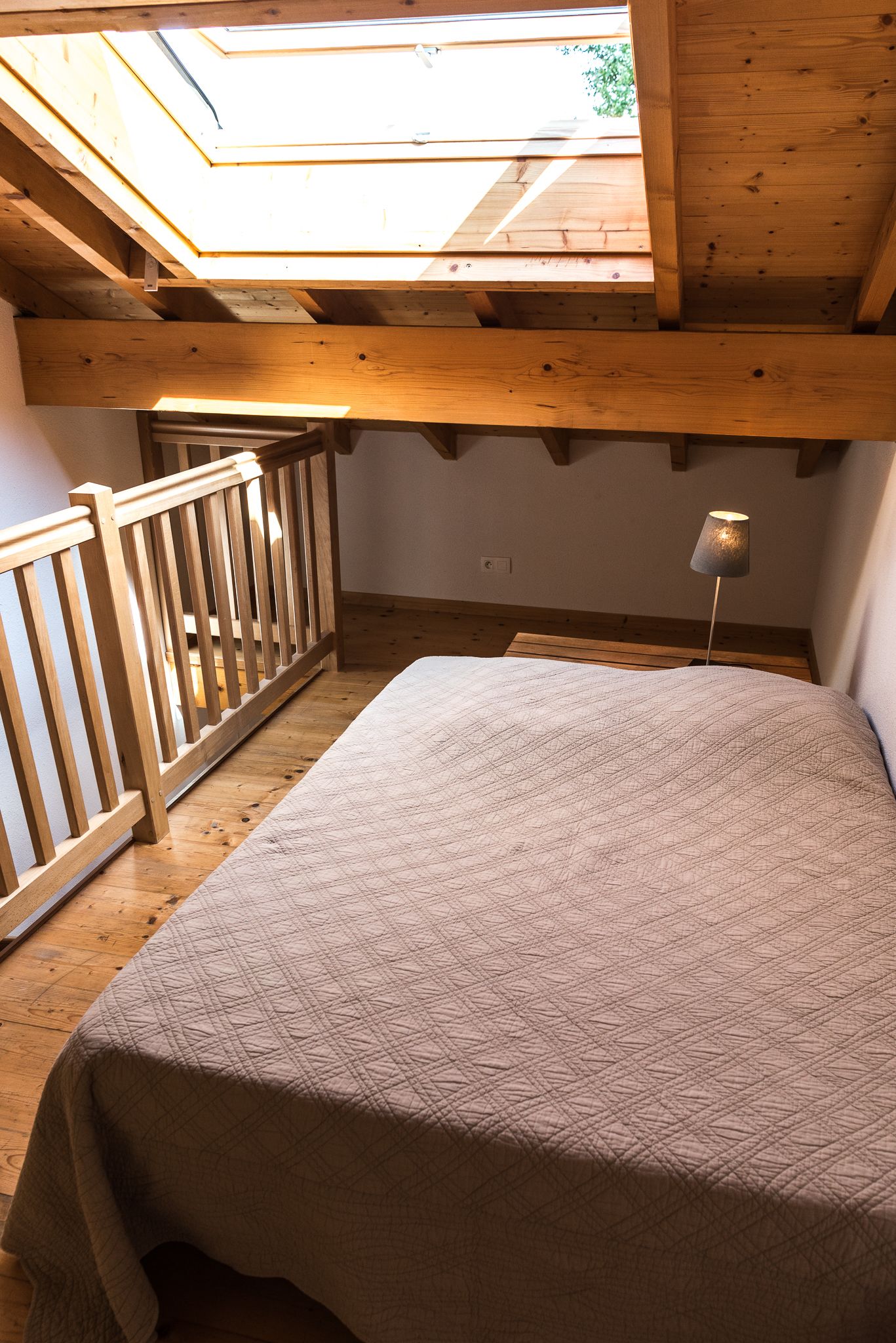 Une chambre avec un lit, des poutres en bois au plafond, une lucarne et une table de chevet avec une lampe.