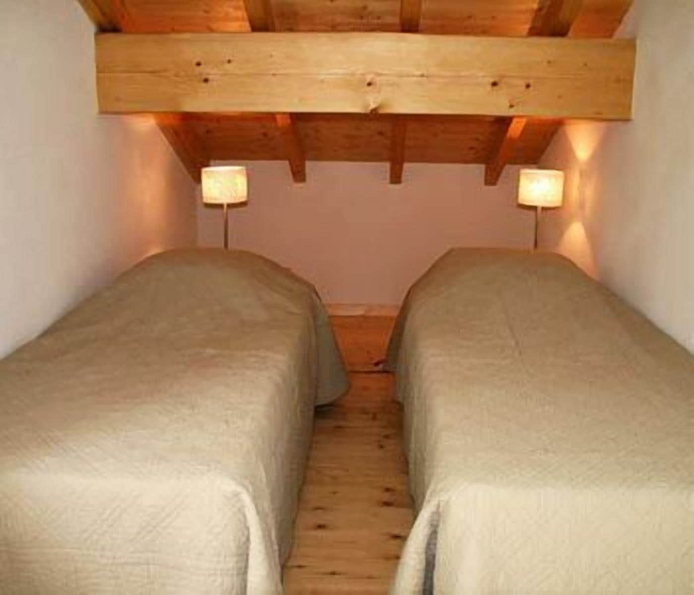 A narrow room with two single beds, wooden beams on the ceiling, and two wall-mounted lamps.