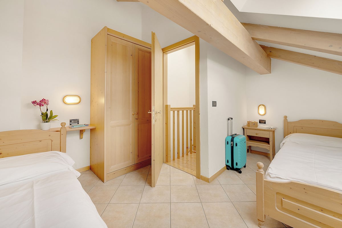 A bedroom with two wooden beds, a wooden wardrobe, a small bedside table with a lamp, and a suitcase on the floor.