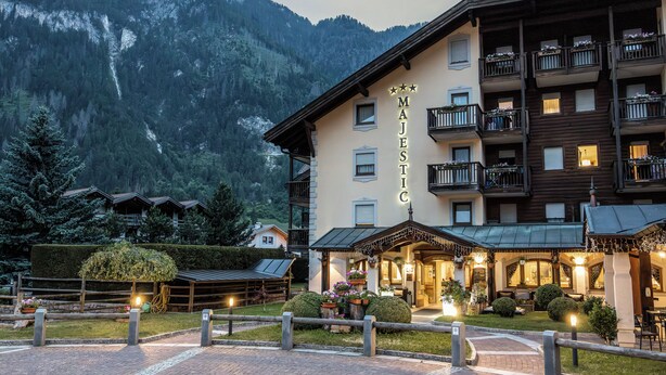 A hotel with a mountain backdrop, multiple balconies, and a well-lit entrance area. - Aparthotel Majestic (Predazzo)