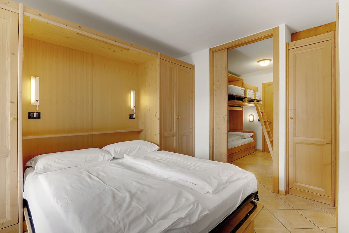 A bedroom with a large bed, wooden wardrobe, and a bunk bed in the background.