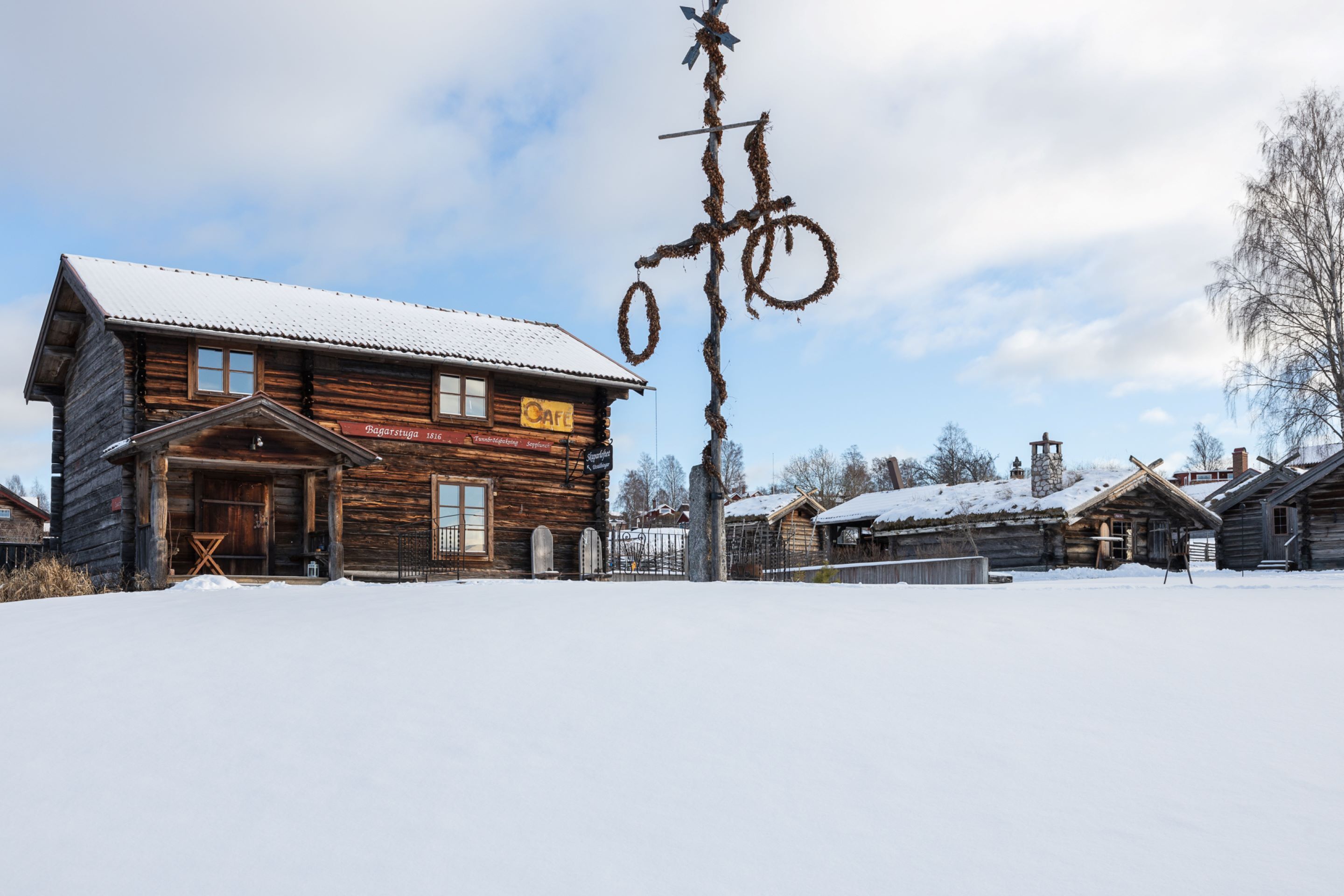 A rustic wooden building with a snow-covered roof, a sign indicating it is a cafe, and a bicycle sculpture in the background.
