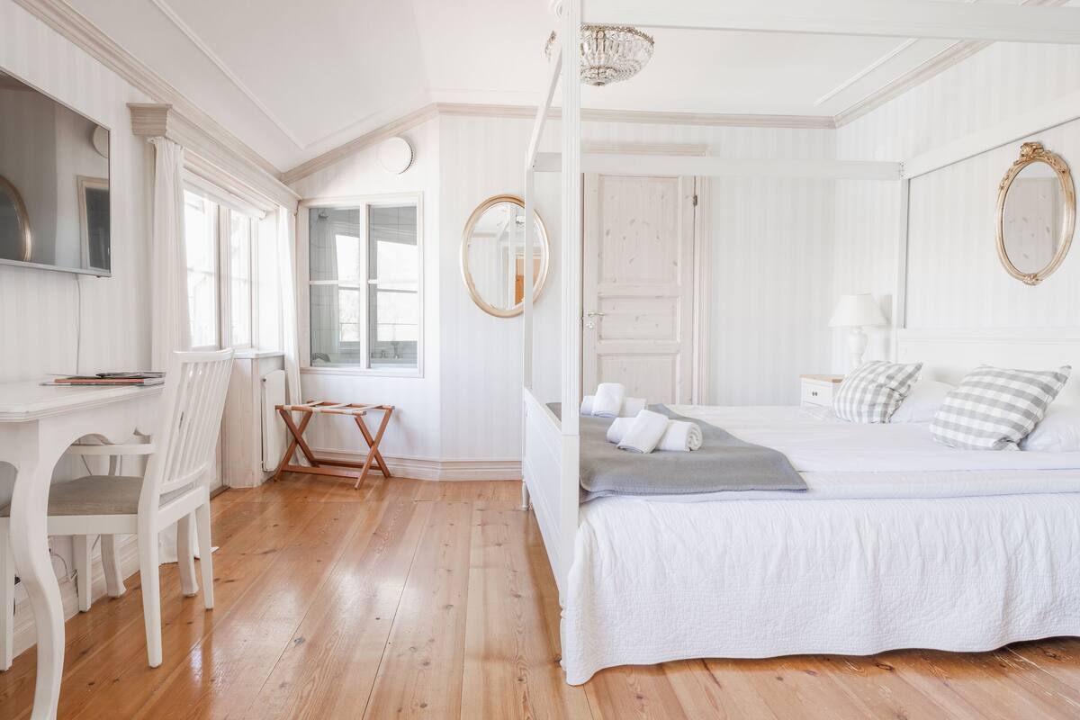 A bedroom with a four-poster bed, a desk with a chair, a mirror, and a window.