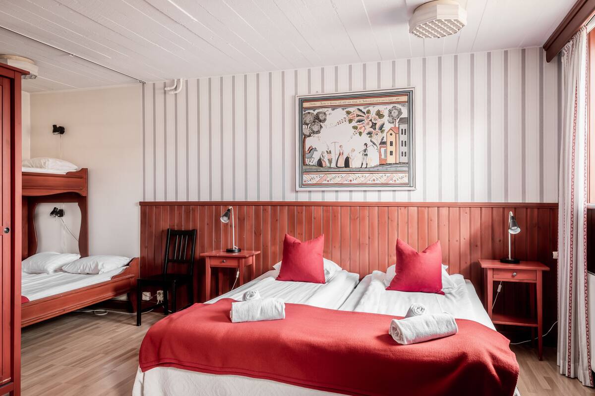A hotel room with a large bed, a smaller bed, a wooden headboard, a nightstand with a lamp, a framed picture on the wall, and a window with curtains.