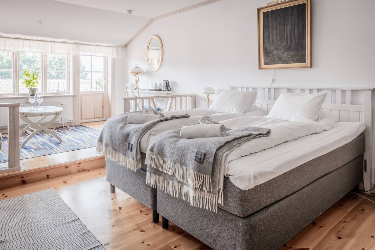 A bedroom with a large bed, a bedside table, a chair, a window with curtains, and a painting on the wall.