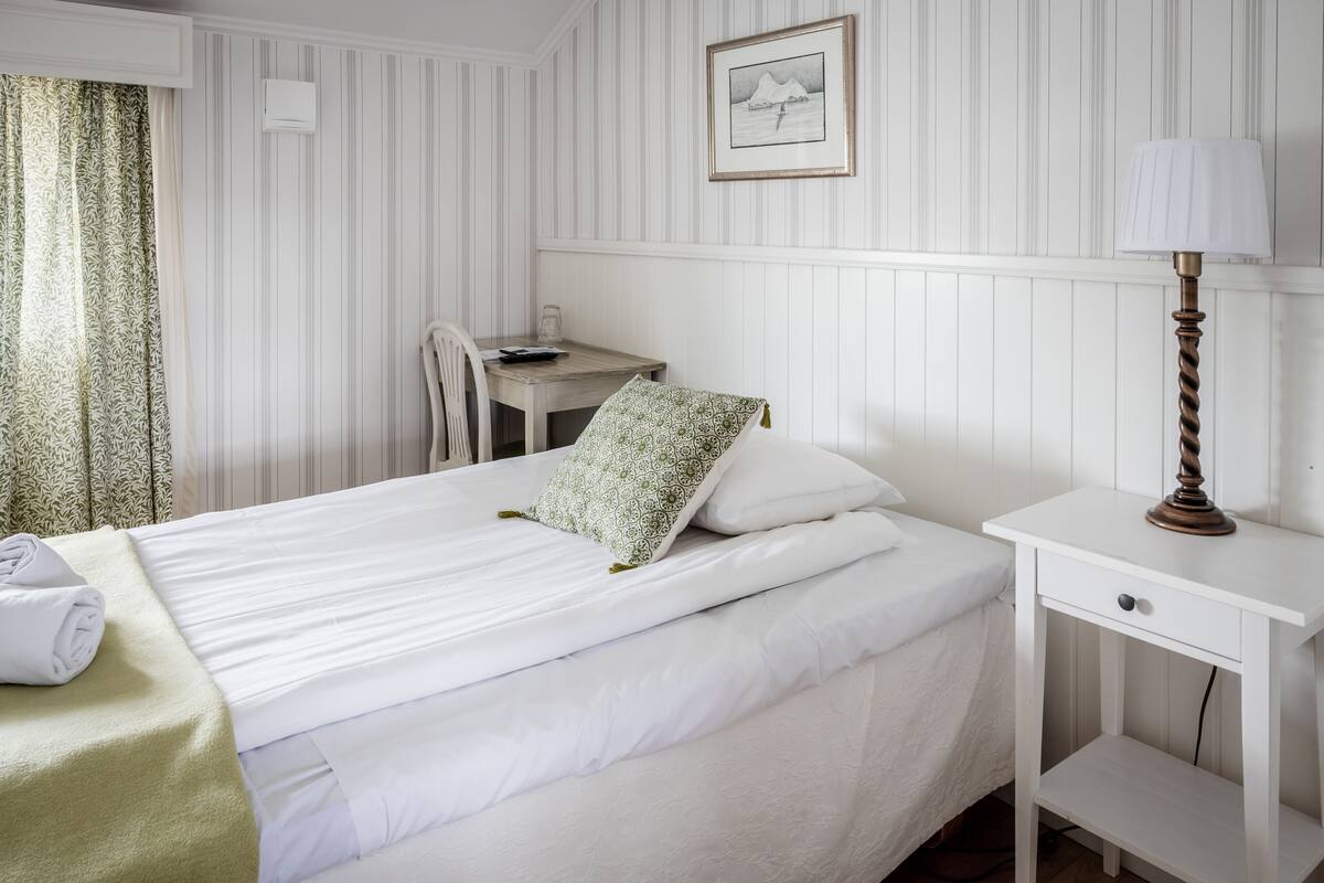 A neatly made bed with pillows, a bedside table with a lamp, a chair, and a desk with a remote control.