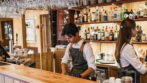 A bar with wooden shelves stocked with various liquor bottles, a bartender preparing drinks, and a customer standing at the counter. - Klockargården Hotell (Tallberg)
