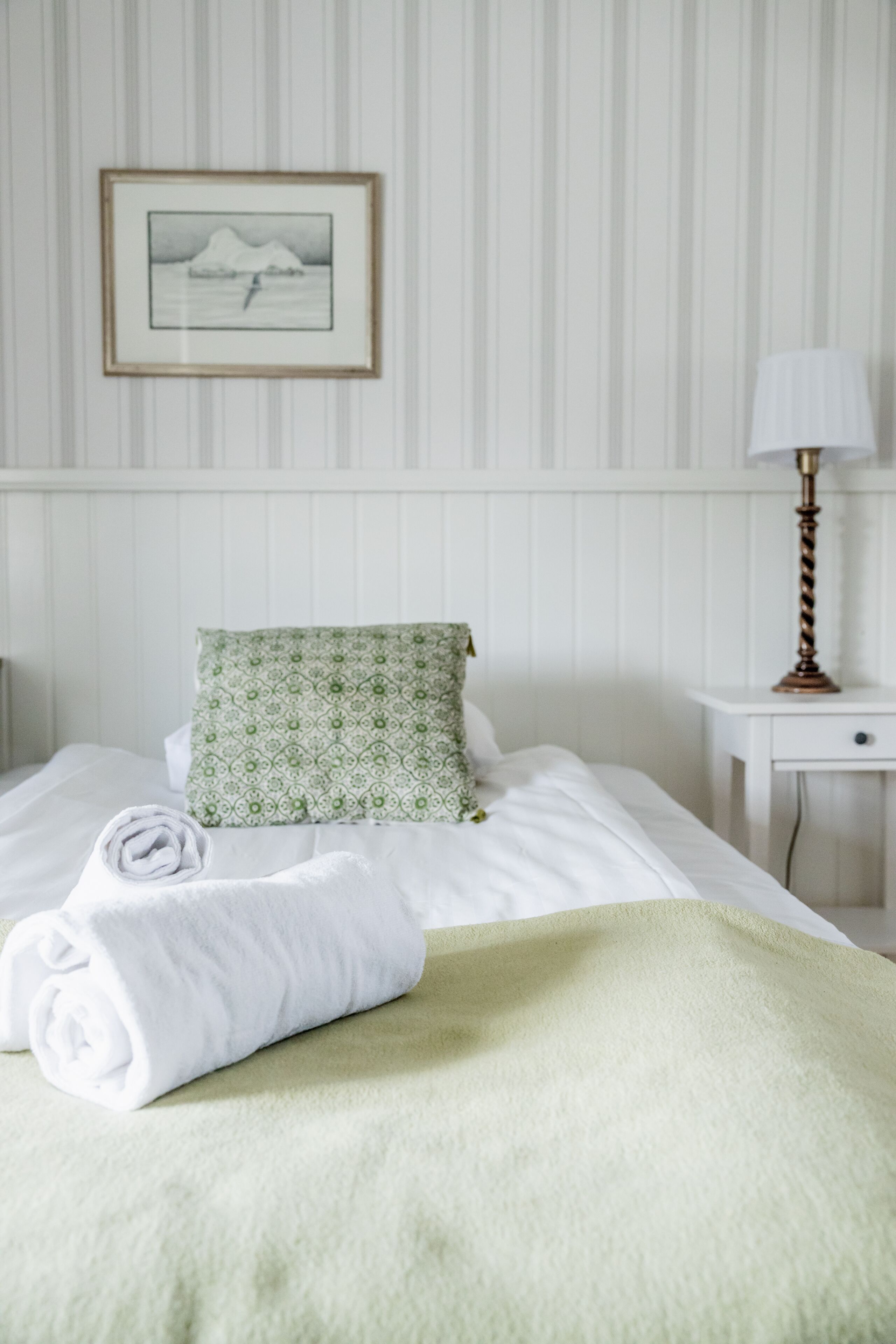 A neatly made bed with a green pillow and a rolled white towel, a framed picture on the wall, and a bedside lamp on a small table.