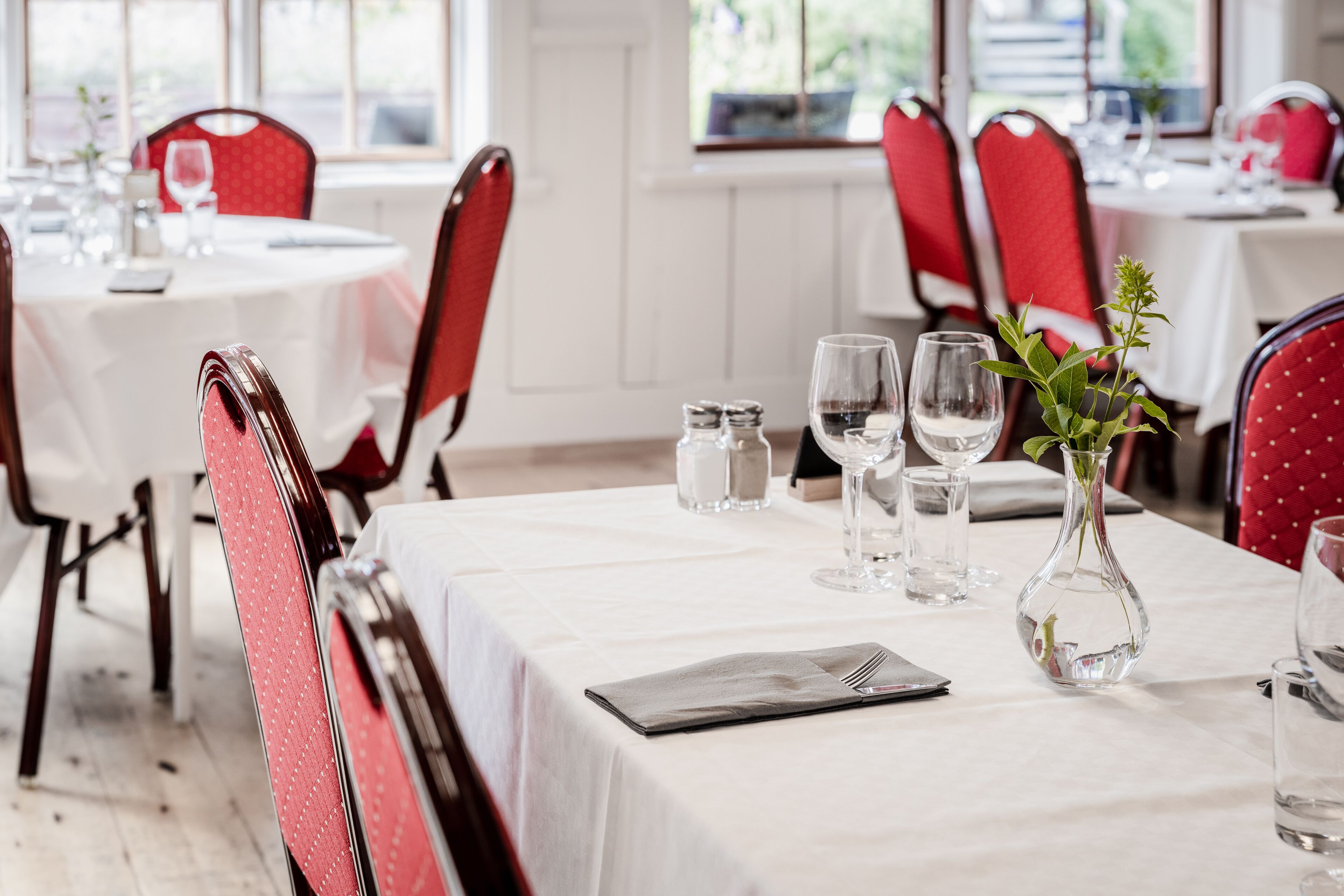 Un comedor con mesas redondas, sillas tapizadas en rojo y manteles blancos.
