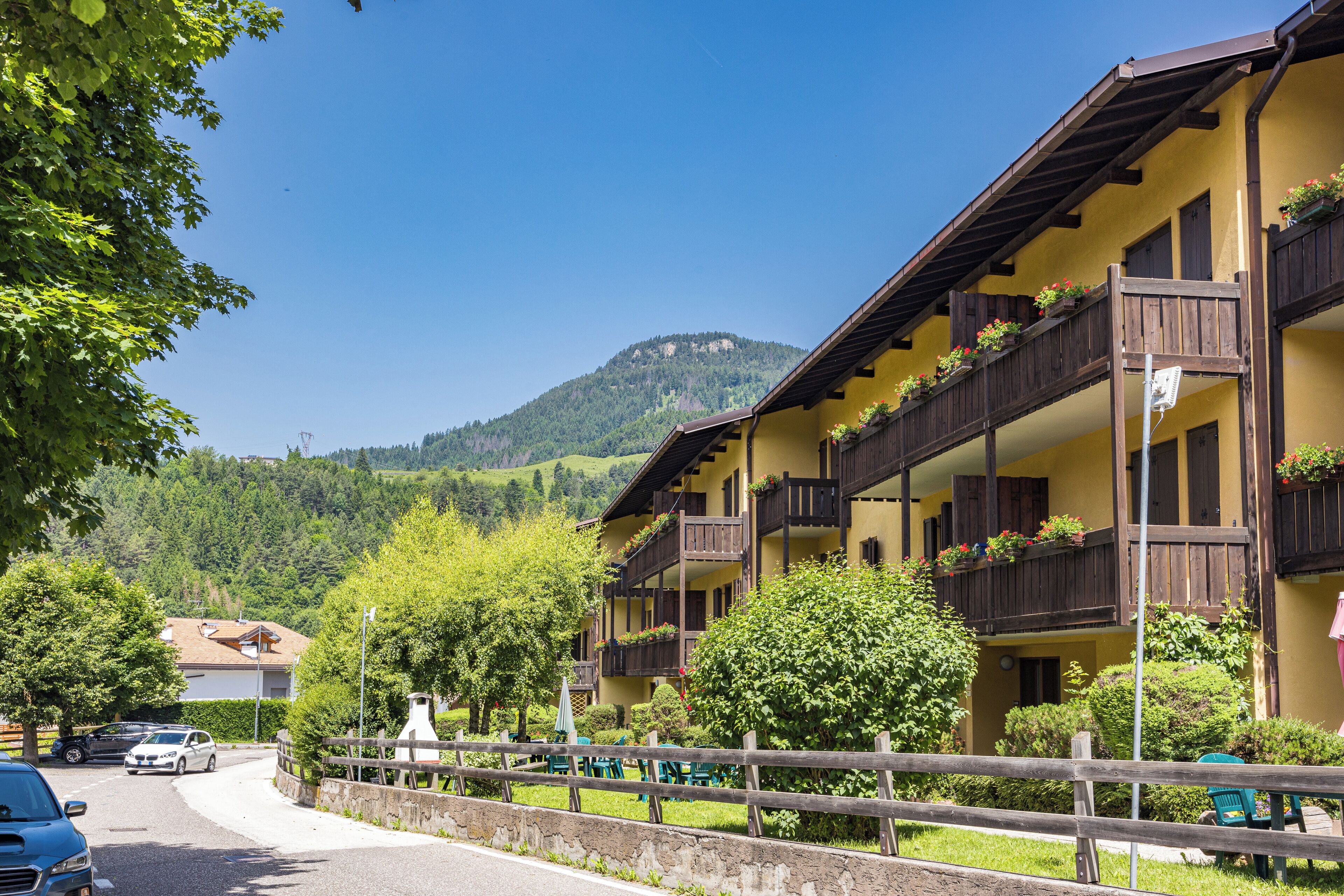 A hotel with balconies adorned with flowers, a mountain backdrop, and a paved road in the foreground.