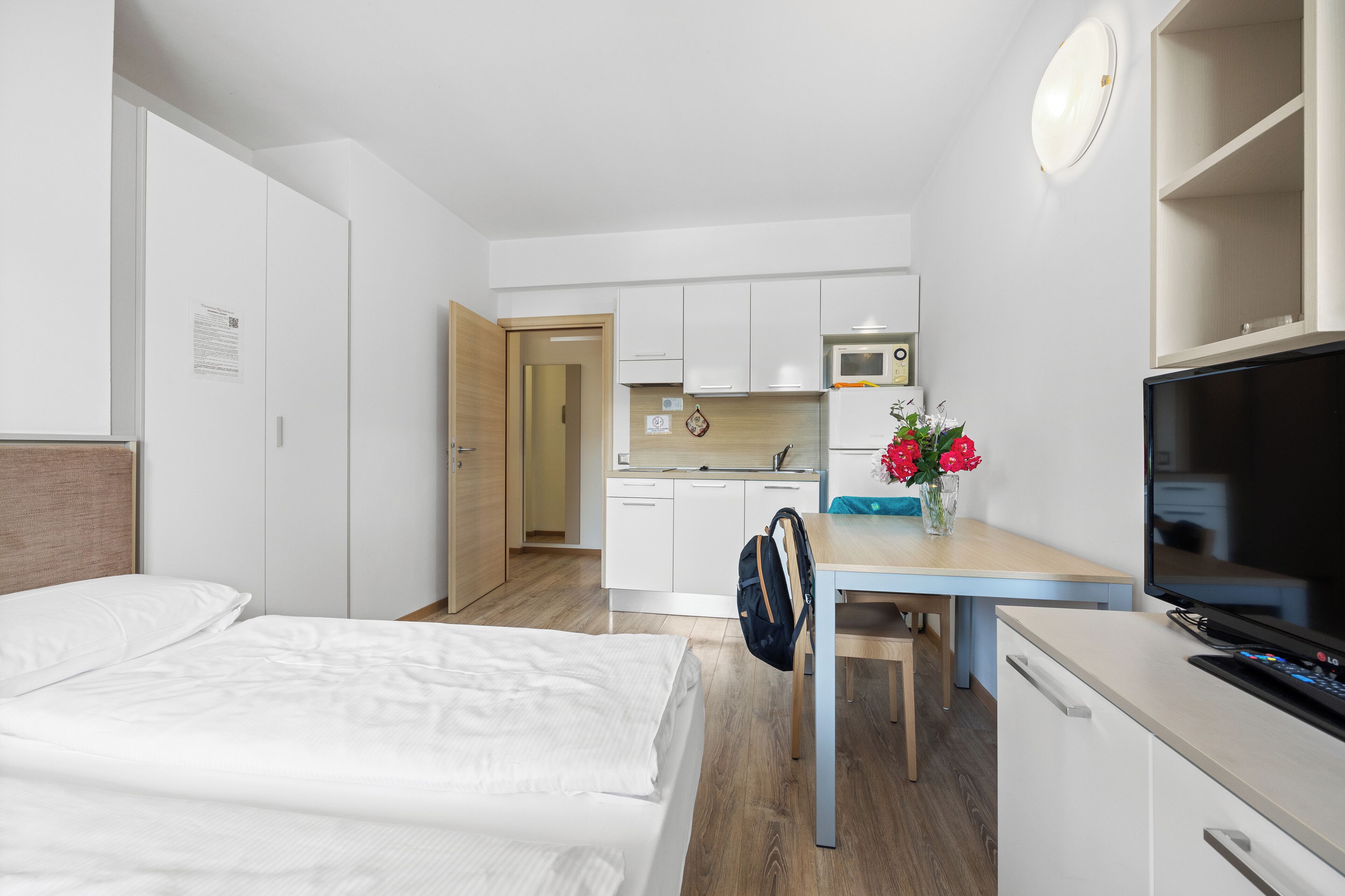 Quarto de hotel com cama, escrivaninha, televisão, micro-ondas e cozinha compacta.
