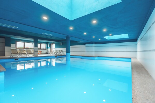 An indoor swimming pool with clear water, a sunroof, and seating areas.
