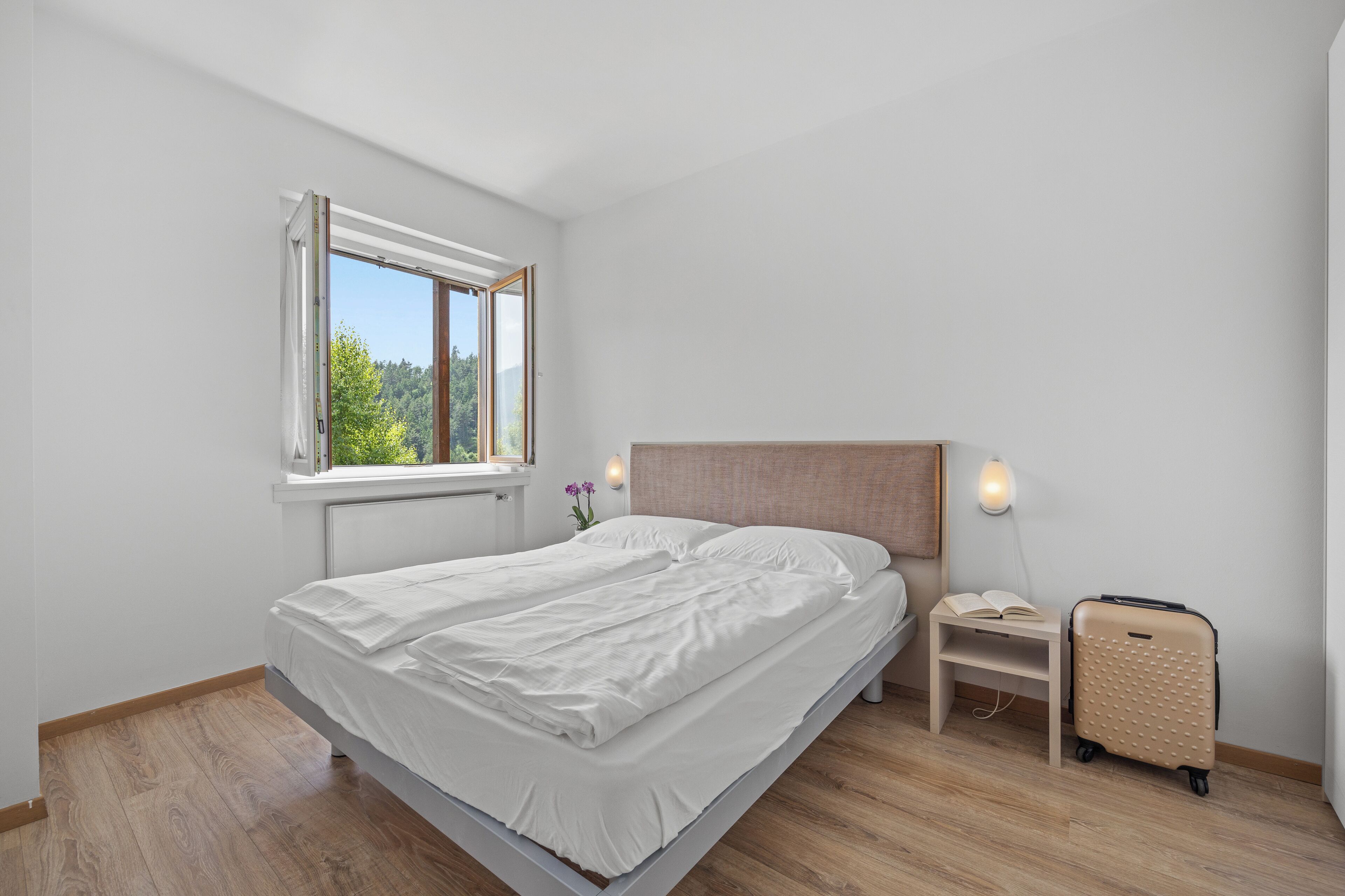 Un dormitorio con cama, ventana, mesita de noche con lámpara y maleta.