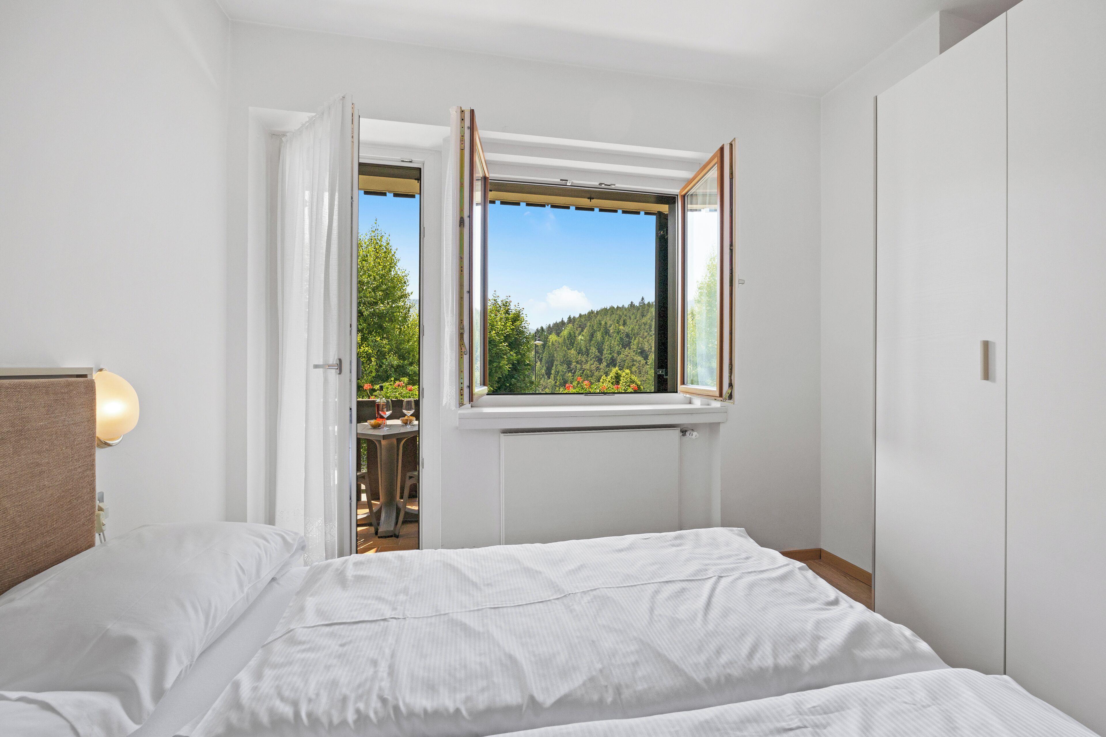 A bedroom with a bed, a window with an open view of a forest, and a door leading to a balcony.