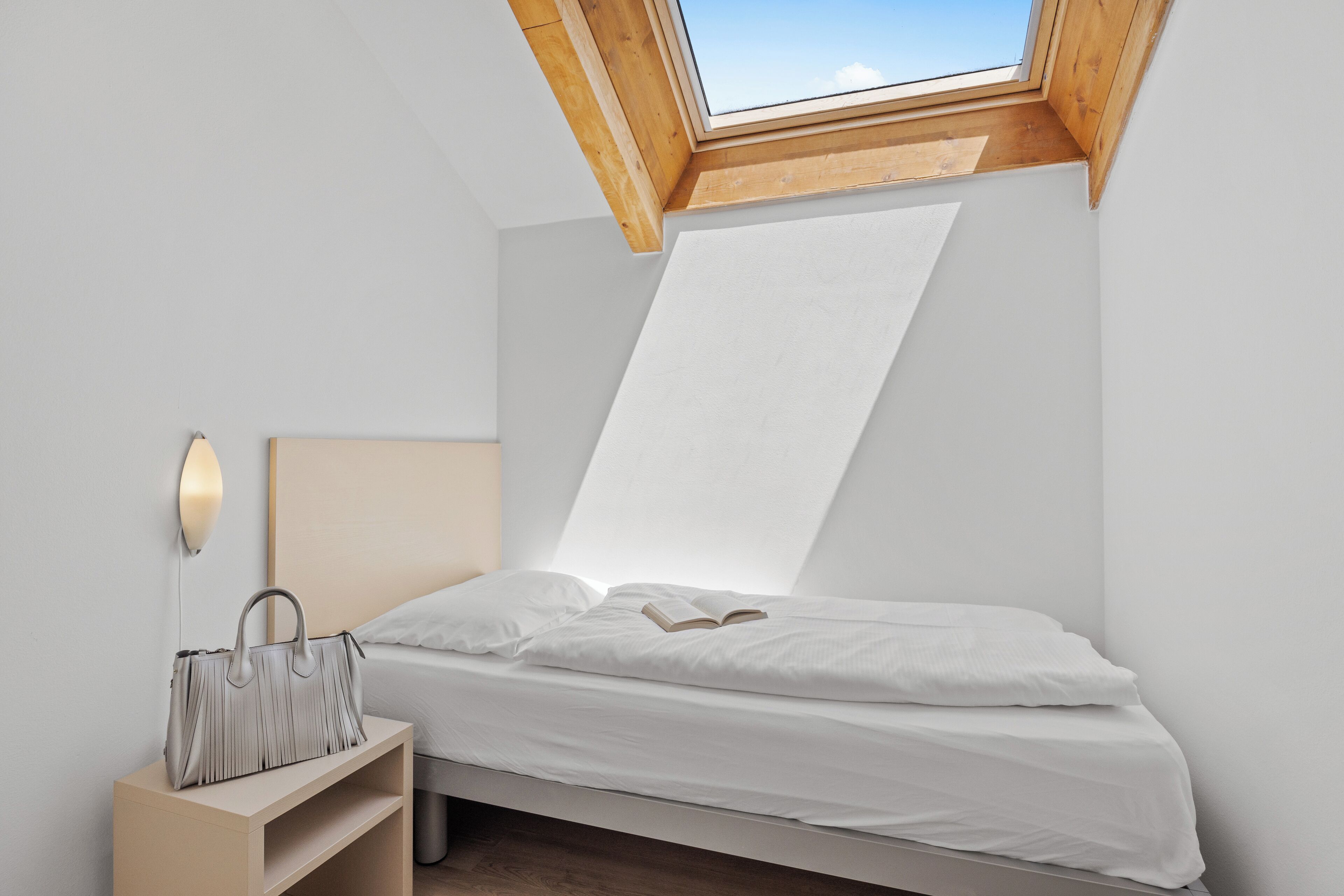 Un dormitorio con tragaluz, cama, mesita de noche y lámpara de pared.