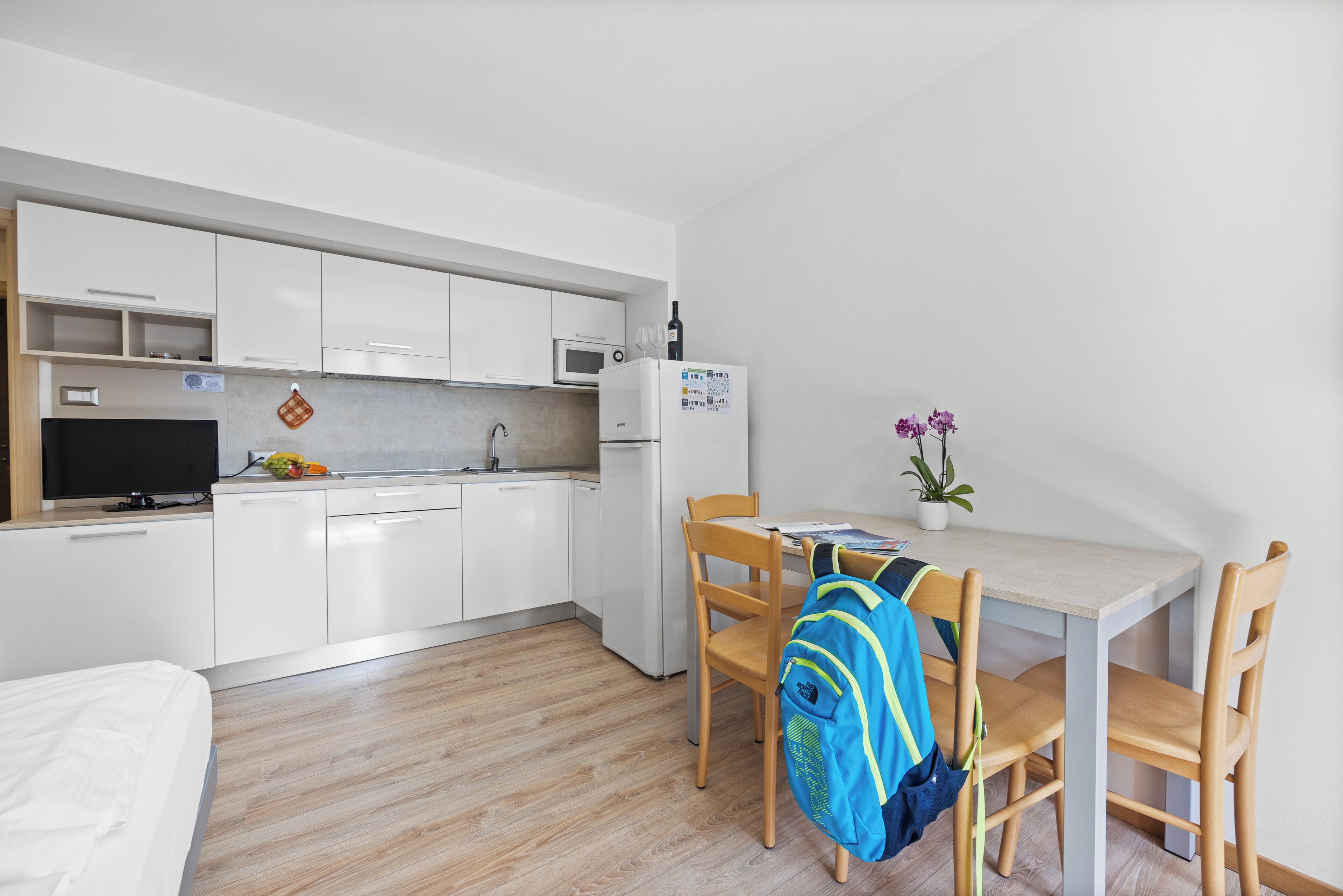 Una cocina compacta con armarios blancos, un microondas y un refrigerador. Hay una mesa de comedor con dos sillas y una mochila sobre una de las sillas. Una pequeña planta en maceta está sobre la mesa.