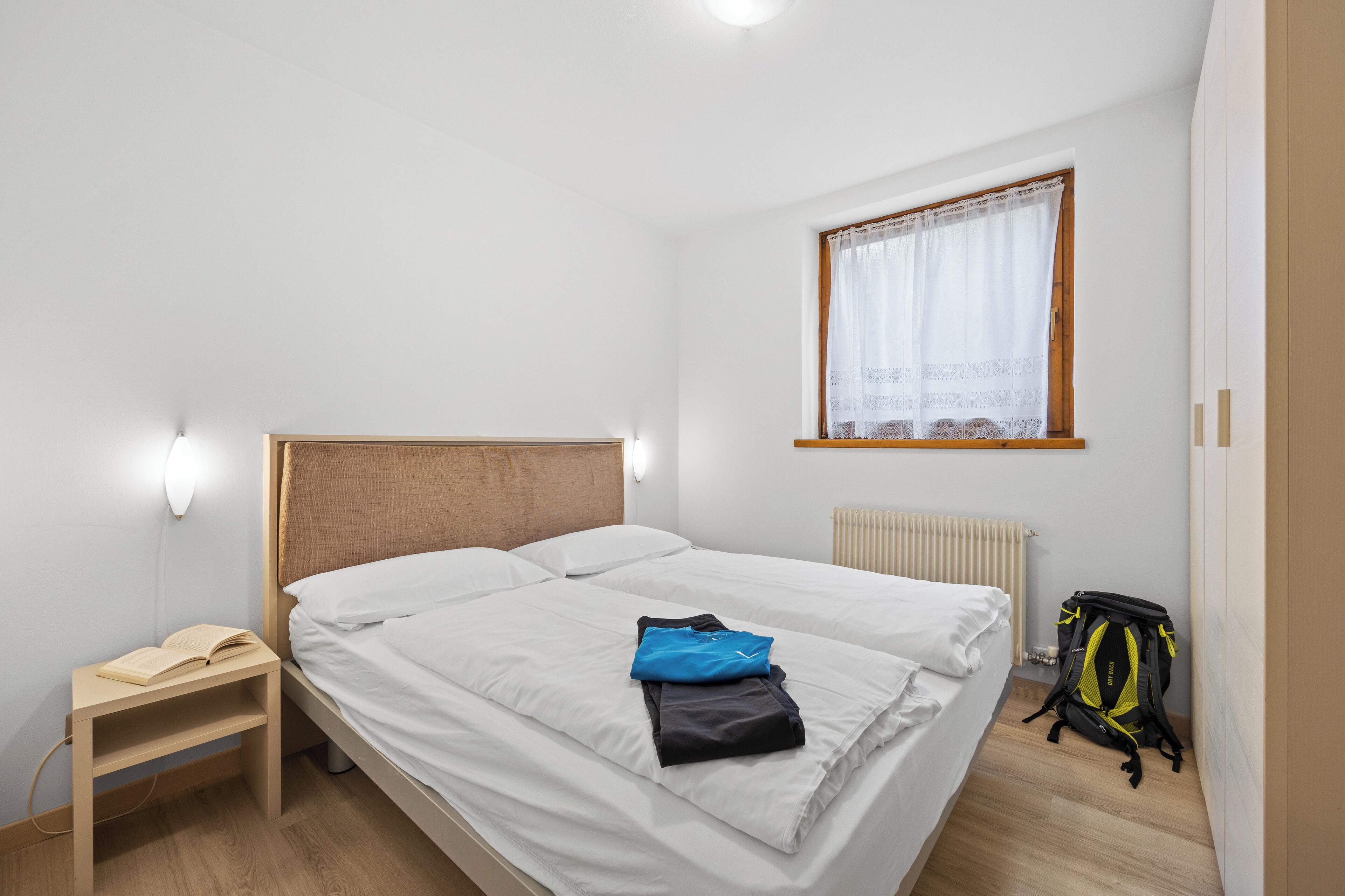 A neatly made bed with white linens, a bedside table with a book, a window with sheer curtains, and a backpack on the floor.