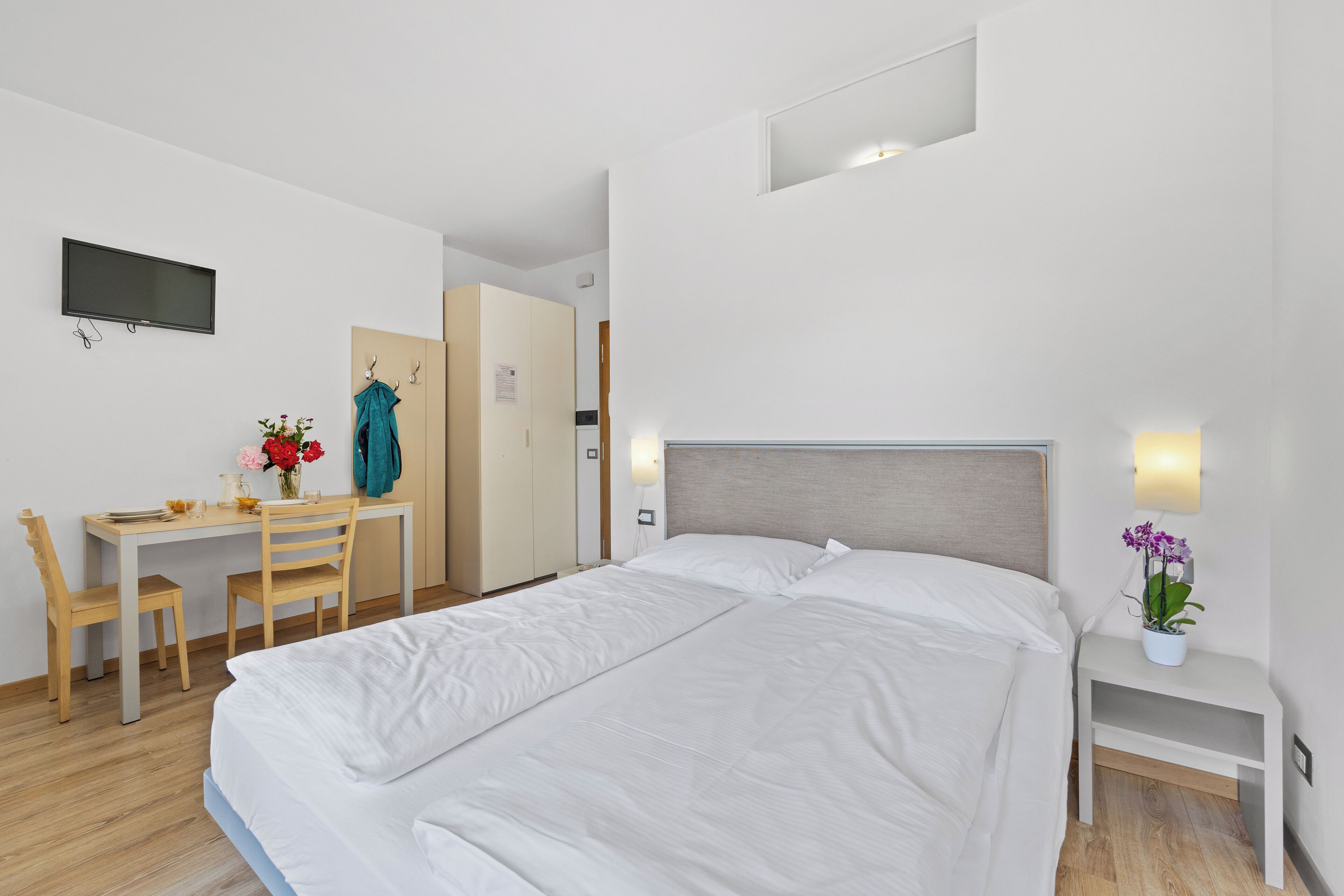 Quarto de hotel com cama, escrivaninha, cadeira, televisão e mesa de cabeceira com abajur e flores.