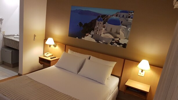 Una habitación de hotel con cama, dos lámparas de noche, una mesita con teléfono y un cuadro colgado en la pared que muestra un pueblo costero.