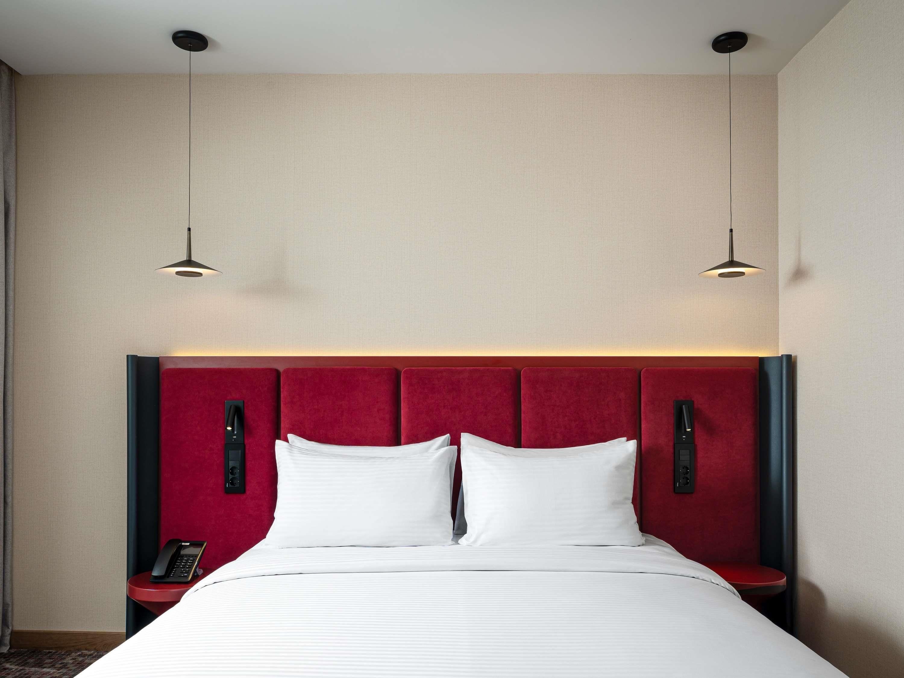 Una habitación de hotel con una cama grande, dos mesitas de noche, un cabecero rojo y dos lámparas colgantes.