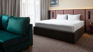 Una habitación de hotel con una cama, un sofá verde, una alfombra estampada y un ventanal con cortinas.