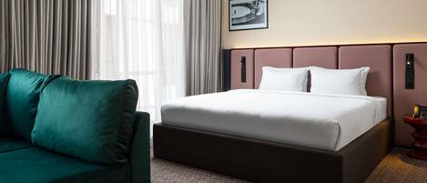 Een hotelkamer met een bed, een groene bank, een kamerbreed tapijt met patroon, en een groot raam met gordijnen.