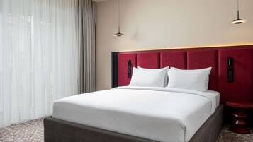 Habitación de hotel con una cama grande, cabecera roja, mesita de noche y ventana con cortinas traslúcidas.