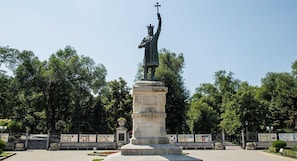 Una estatua de una figura sosteniendo una cruz sobre un pedestal en un parque con árboles y una valla al fondo.