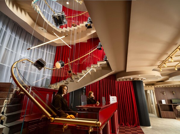 A grand staircase with red curtains, a piano, and a reception desk in a luxurious lobby.