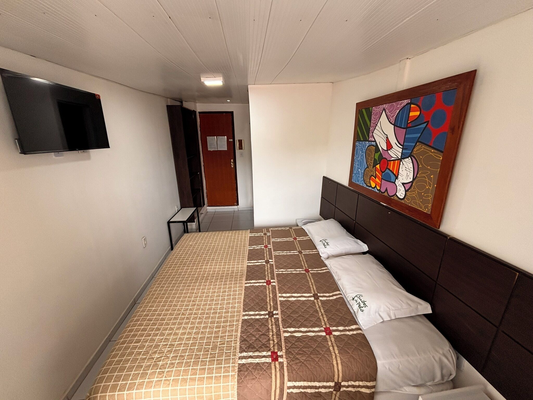 Une chambre d’hôtel avec un lit, un téléviseur à écran plat, un tableau coloré et une porte donnant sur un couloir.