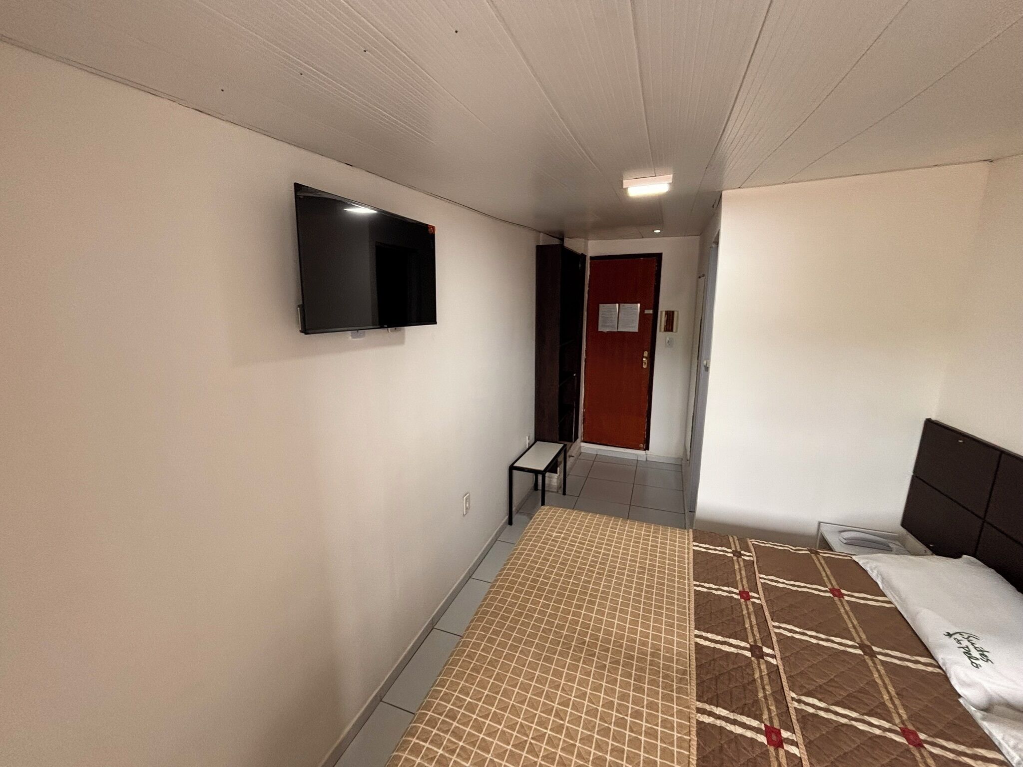 A hotel room with a bed, a TV mounted on the wall, a small table, and a door leading to another room.