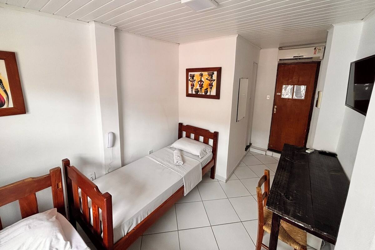 Een enkel bed met een houten hoofdbord, een nachtkastje, een stoel, een bureau en een wandairconditioner.